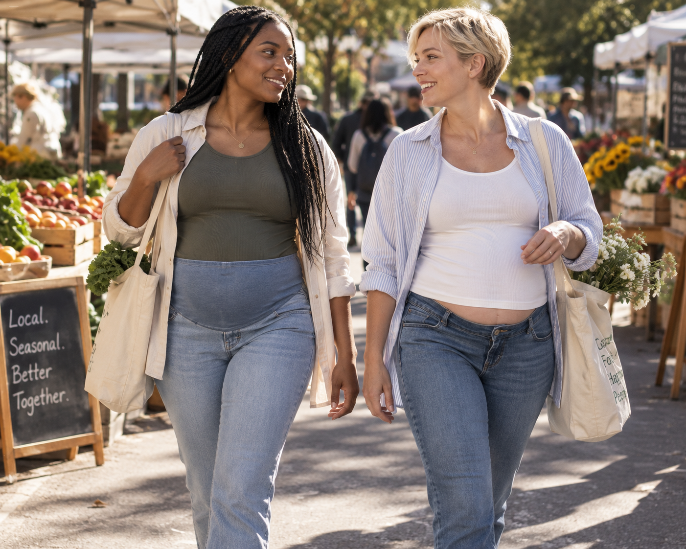 To gravide kvinner går sammen på et utendørs marked, en i jeans over magen og en i jeans under magen.